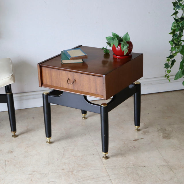 British mid-century vintage teak bedside cabinet with black tapered legs and gold feet, drawer with wood grain detail,.