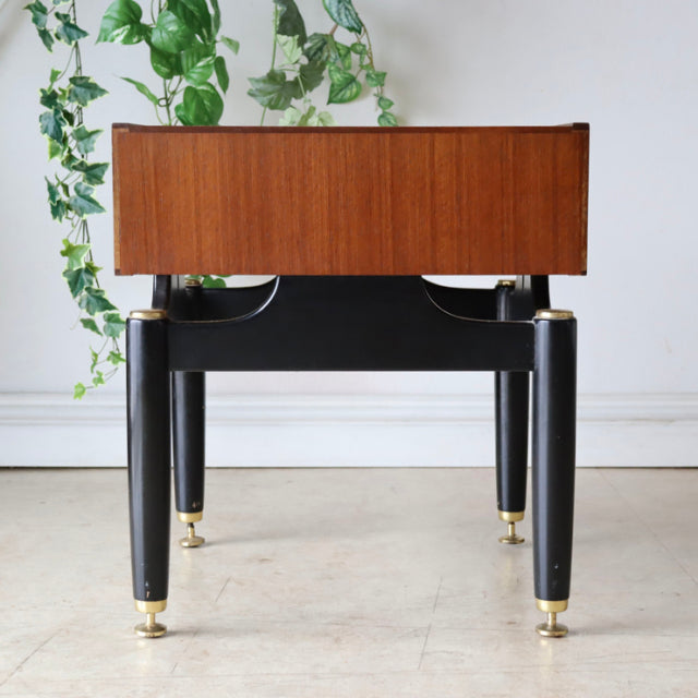 Vintage British mid-century teak bedside cabinet with black tapered legs and brass feet, sleek rectangular form, visible.