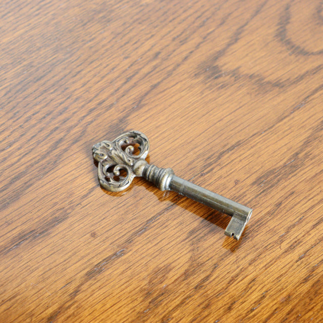 Ornate vintage metal key with intricate scrollwork resting on warm teak wood grain surface, evocative of British mid-century .