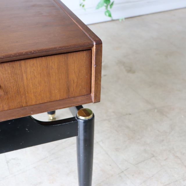 Vintage British mid-century teak bedside cabinet with black tapered legs and gold accents, sleek clean lines, smooth wood.