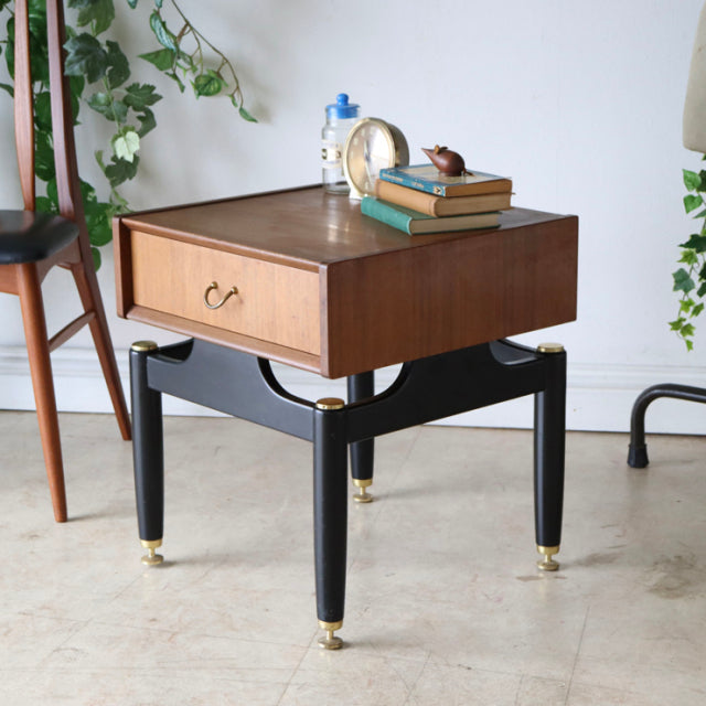 Mid-century British G-Plan teak bedside cabinet with black legs, gold accents, wood grain top, and drawer handle.