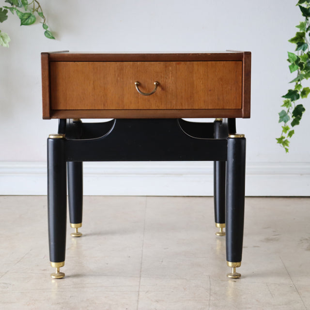 Vintage British mid-century teak bedside cabinet with rich wood grain, black tapered legs, and gold accents.