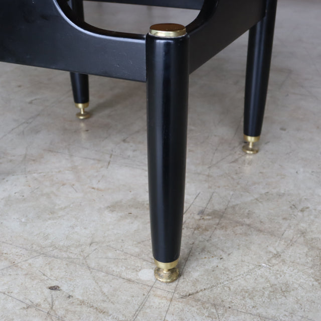 Mid-century British G-Plan teak bedside cabinet leg in black with brass cap and foot on concrete floor.