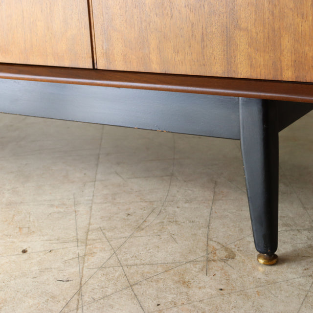 Close-up of mid-century British G-Plan sideboard leg in black with brass cap, vintage teak wood grain visible above.