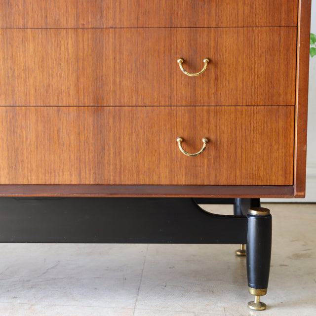 Vintage British G-Plan mid-century teak chest with wood grain, black tapered legs, brass handles, and brass feet accents.