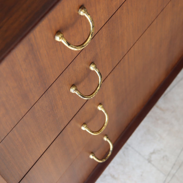 Vintage British G-plan teak four-drawer chest with curved brass handles, mid-century design, smooth wood grain, slender legs.