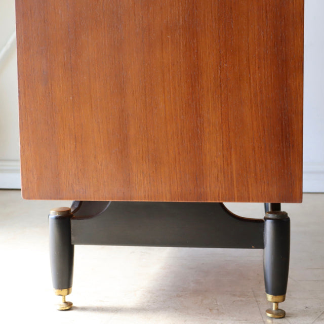 Mid-century British G-Plan teak chest close-up showing rich wood grain and tapered black legs with brass caps against white.