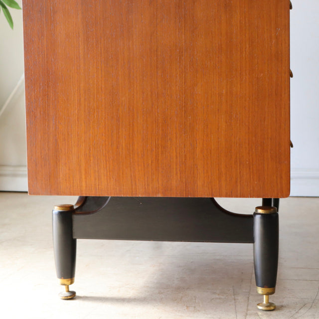 Mid-century British G-Plan teak chest side view showing smooth wood grain and black tapered legs with brass feet.
