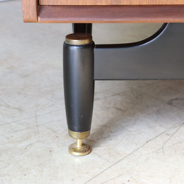 Close-up of vintage British G-Plan mid-century teak sideboard leg with black tapered wood and brass foot detail on concrete.