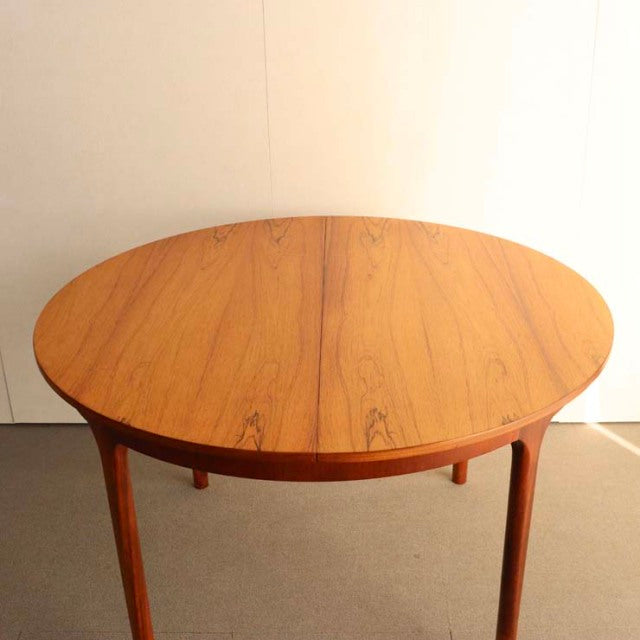 Vintage teak round dining table with smooth wood grain, slender tapered legs, mid-century British-Denmark design.