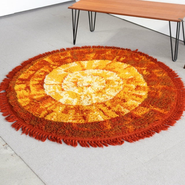 Round vintage mid-century rug in warm orange and yellow tones with fringed edges, featuring a spiral pattern on a light.