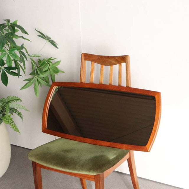 Mid-century British vintage teak mirror with rich wood grain and curved rectangular frame, leaning on green velvet seat chair.
