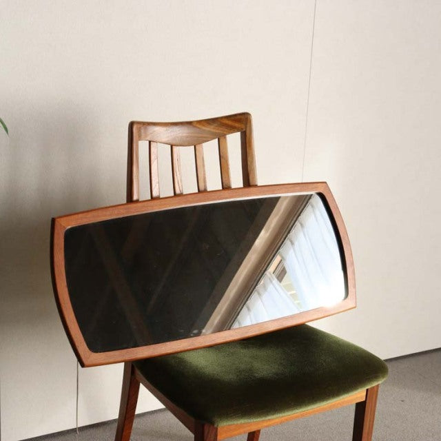 Mid-century British vintage teak mirror with curved rectangular frame and visible wood grain, resting on green velvet chair.