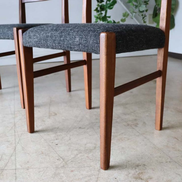 Set of four mid-century British vintage teak dining chairs with dark textured fabric seats and sleek, tapered legs showing.