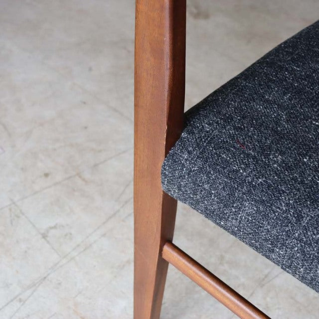 Close-up of vintage British mid-century teak dining chair leg with dark textured fabric seat and smooth wood grain finish.