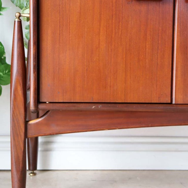 Vintage teak sideboard corner showing rich wood grain, tapered British mid-century legs with brass caps, smooth front panel.