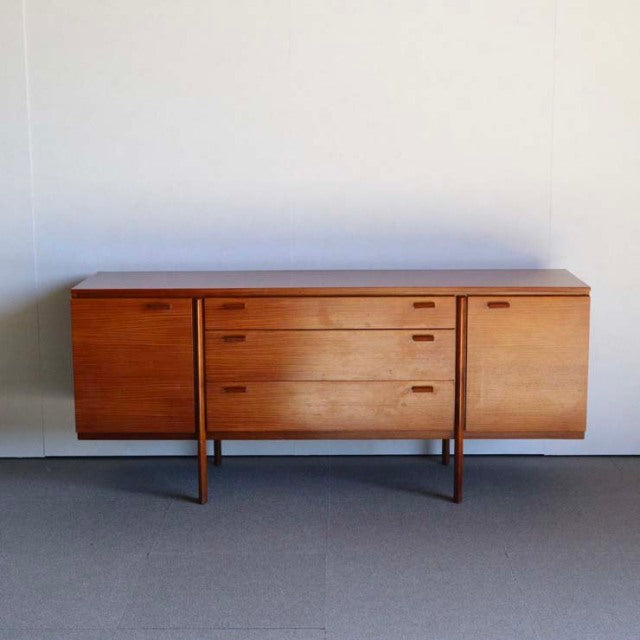 Mid-century British vintage teak sideboard with rich wood grain, three central drawers, and two side cabinets on slender legs.