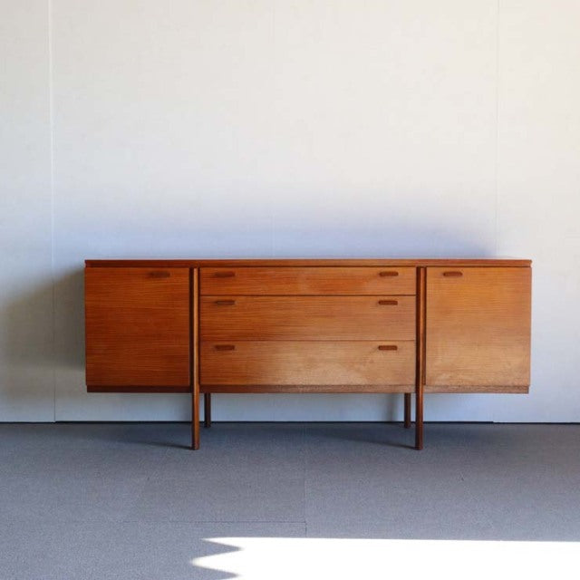 Mid-century British vintage teak sideboard with rich wood grain, three central drawers, and two side cabinets on slender legs.