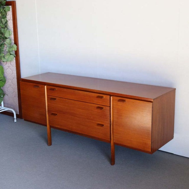 Mid-century British vintage teak sideboard with smooth wood grain, tapered legs, and three drawers flanked by cupboards.