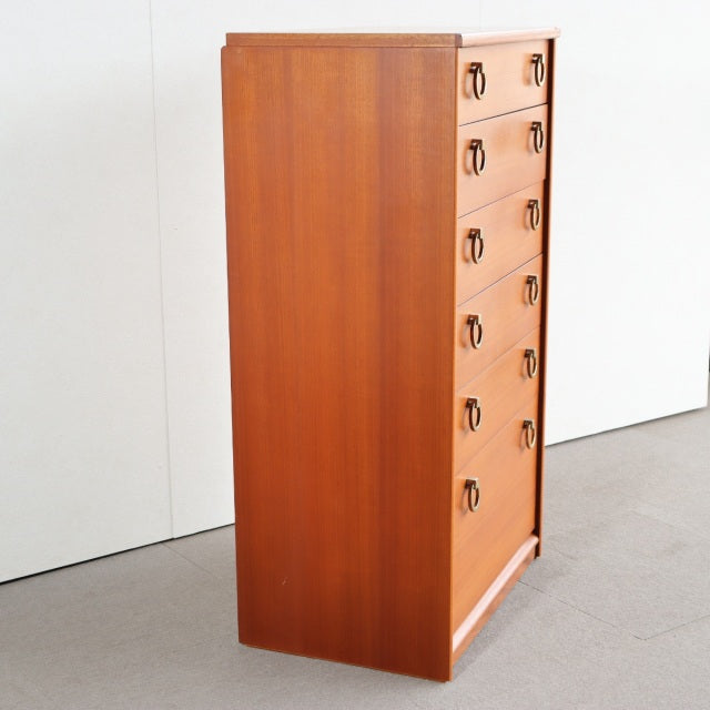 Vintage British mid-century teak chest with seven drawers, brass ring handles, sleek wood grain, and slender legs.