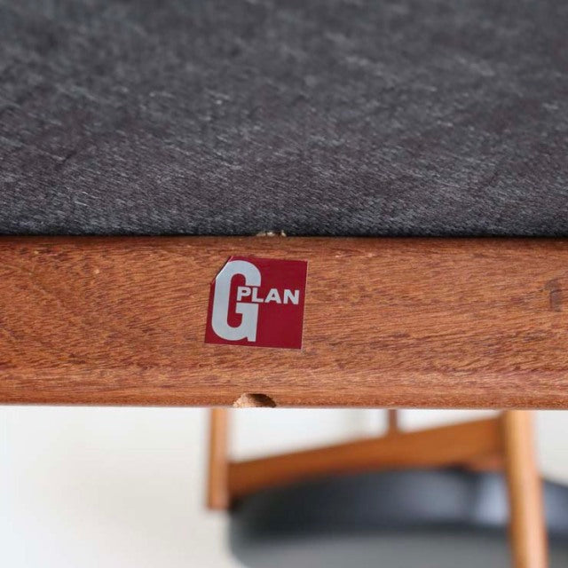 Close-up of mid-century G-plan teak chair frame with visible wood grain and red G-plan label under gray fabric seat.