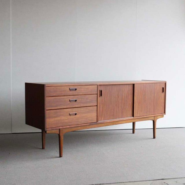 Mid-century British teak sideboard by Nathan with three drawers, sliding doors, slender tapered legs, and rich wood grain.