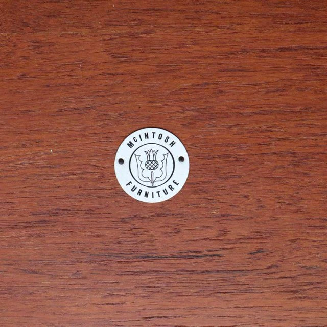 Round white McIntosh Furniture logo medallion on vintage teak wood with rich mid-century British Denmark grain.