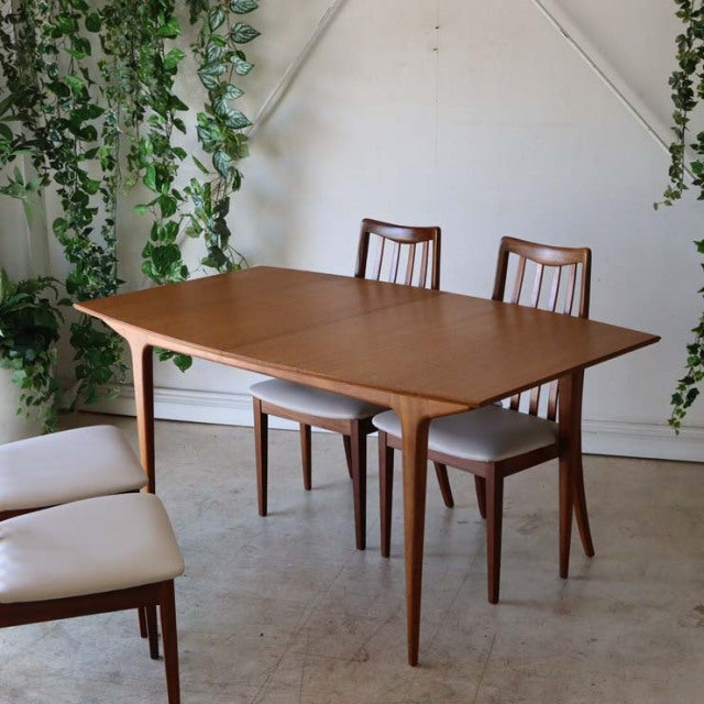 Vintage mid-century British teak dining table with smooth wood grain, tapered legs, paired with matching teak chairs.