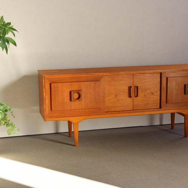 Vintage British mid-century teak sideboard with rich wood grain, square recessed handles, and tapered legs, reflecting.