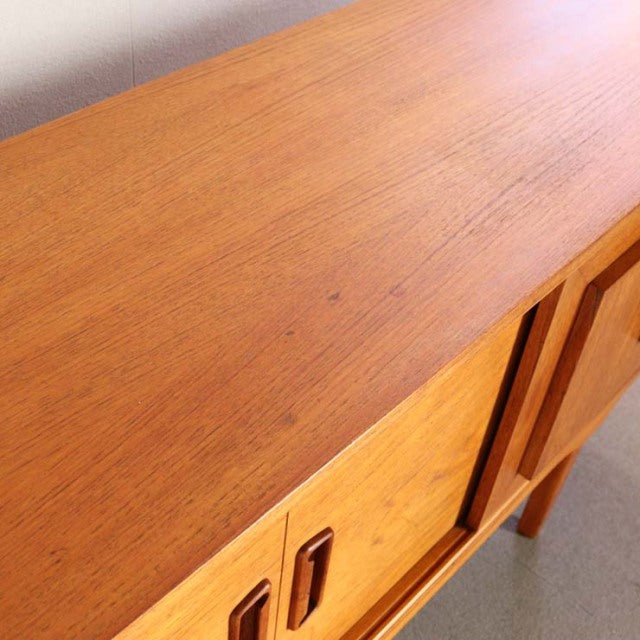 Vintage British mid-century teak sideboard with smooth wood grain, tapered legs, and recessed handles.