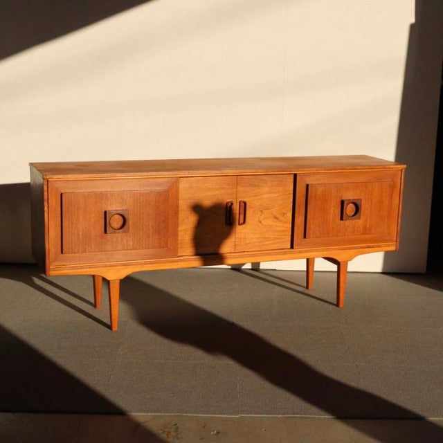 Vintage mid-century British teak sideboard with rich wood grain, three cabinet doors, and tapered legs in warm sunlight.