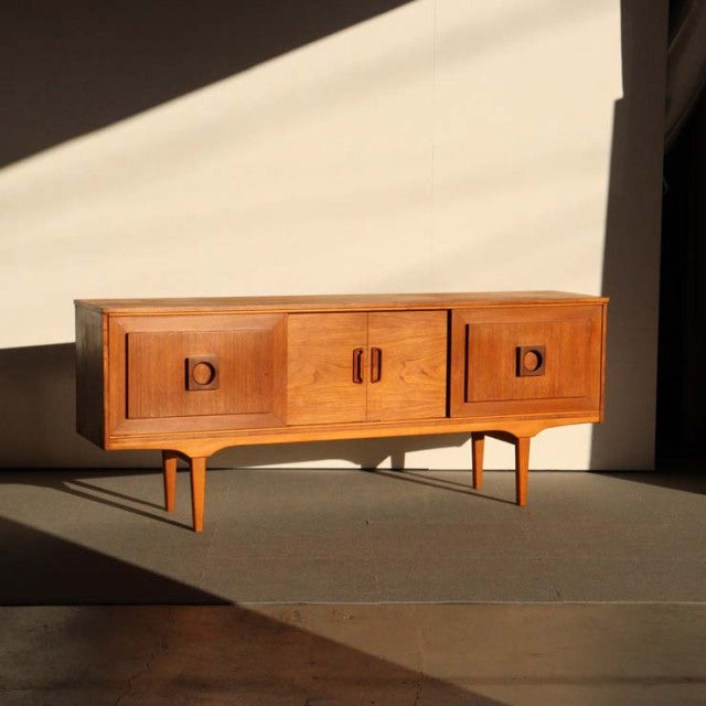 Mid-century British vintage teak sideboard with rich wood grain, tapered legs, and three cabinet doors.
