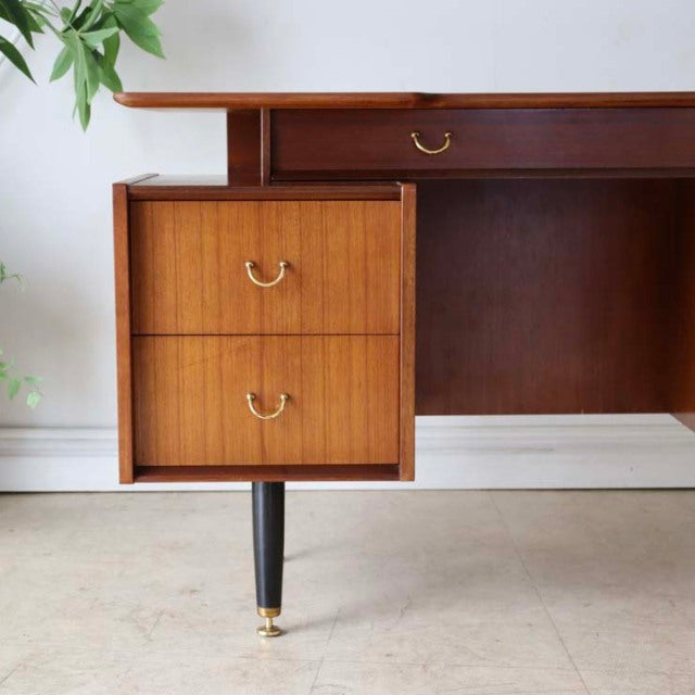 Mid-century British G-PLAN teak desk with visible wood grain, brass handles, and tapered black legs with brass caps.