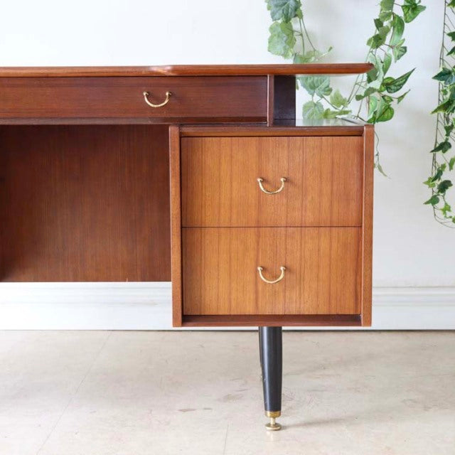 Mid-century British G-PLAN teak desk with rich wood grain, two drawers with brass handles, and slender black tapered legs.