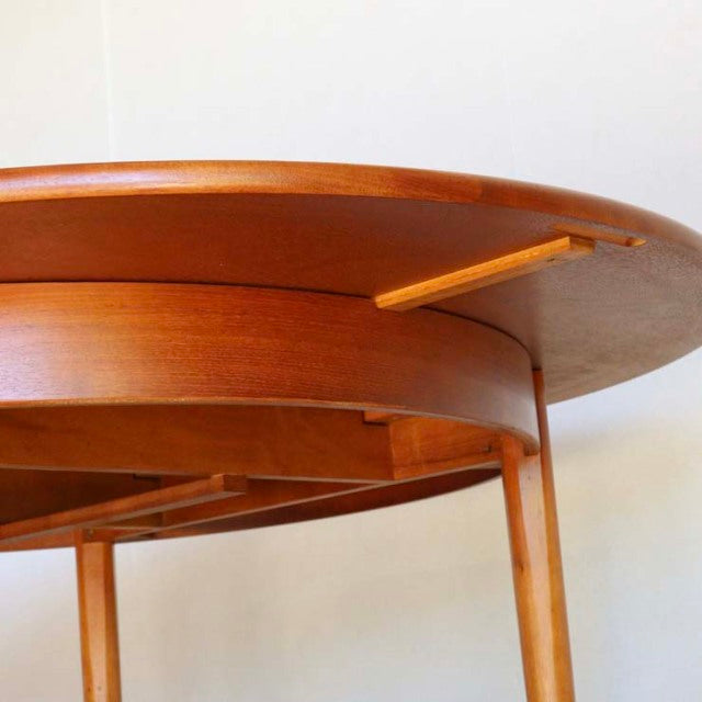 Round teak dining table with mid-century British vintage design, smooth wood grain, slender tapered legs, and subtle.