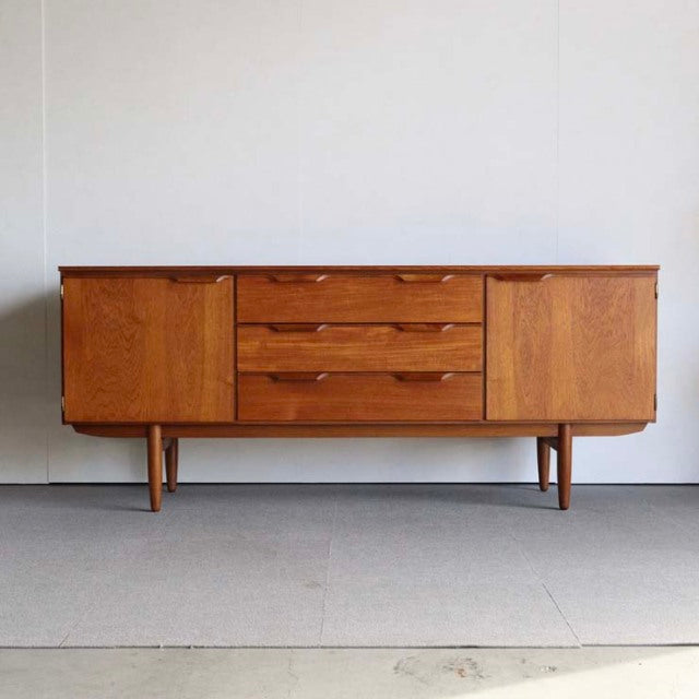 Mid-century vintage British sideboard in teak with rich wood grain, slender tapered legs, and three central drawers flanked.