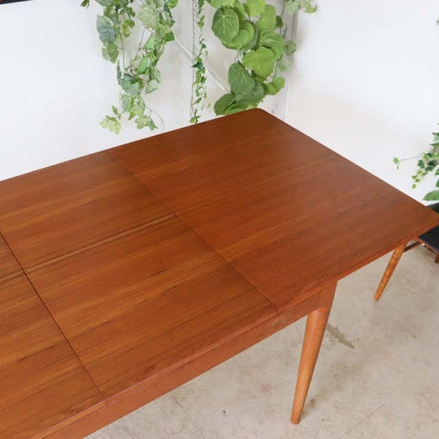 Mid-century vintage Danish teak dining table with rich wood grain and tapered legs against a white wall with green plants.