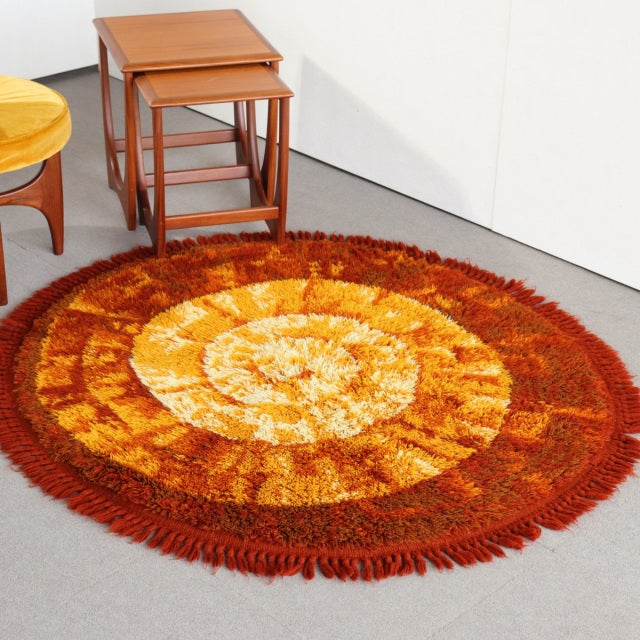 Round vintage mid-century rug with bold orange, yellow, and brown spiral pattern, textured wool fibers, and fringed edges,.