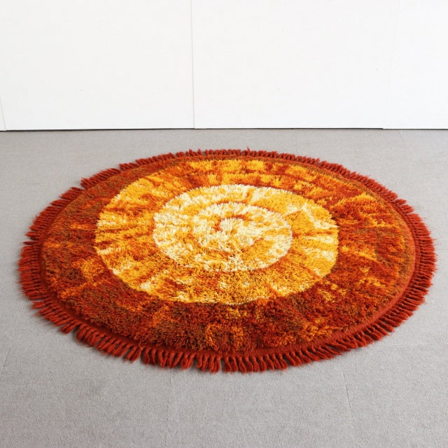 Round vintage shag rug with warm orange, yellow, and brown tones, textured spiral pattern, fringed edges, mid-century.